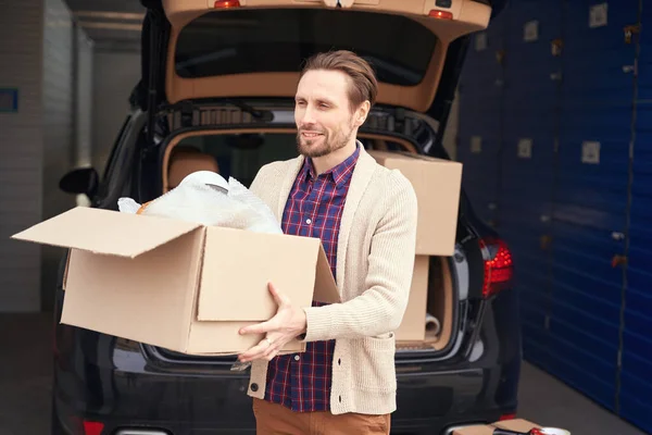 快乐的家伙站在仓库里一辆敞着的汽车行李箱旁边 手里拿着一个大纸板箱 — 图库照片
