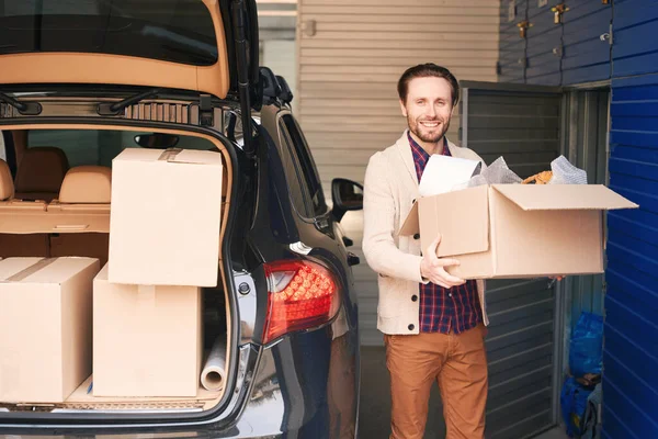 幸せな男は倉庫に箱を持った車のトランクの近くに立っている大きな段ボール箱を持っている — ストック写真
