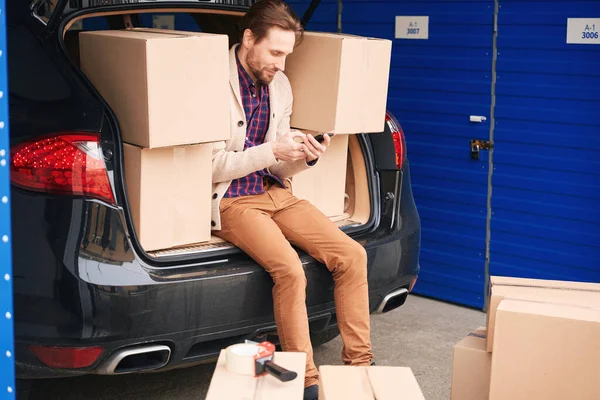 这个年轻人坐在仓库里 坐在汽车后备箱里打电话 装有搬运物品的纸板箱在行李箱里 在汽车旁边 — 图库照片