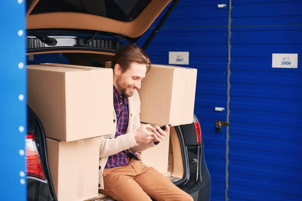 快乐的家伙坐在仓库里 坐在汽车后备箱里打电话 装有搬运物品的纸板箱在行李箱里 — 图库照片