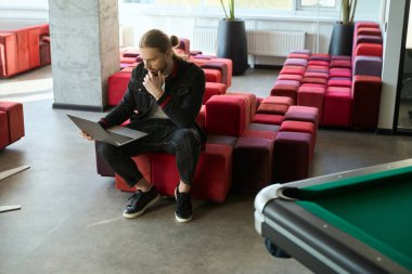 Süslü giysiler içinde beyaz bir adam iş yerinde dizüstü bilgisayarla çalışırken düşünüyor.