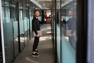 Gülümseyen adam başparmaklarını kaldırırken ve içerideki kameraya bakarken elinde defter bilgisayarı tutuyor.