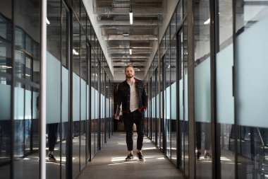 Yakışıklı bir erkek, kameraya bakıyor ve koridorda yürürken elinde bilgisayar tutuyor.