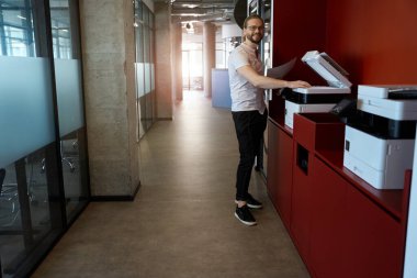 Birlikte çalışma alanında fotokopi makinesi kullanarak notları kopyalarken kameraya bakan süslü elbiseli gülen adam.