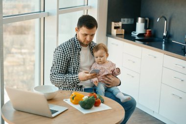 Ev elbiseli bir erkek mutfak masasında oturuyor, kollarında sevimli bir bebek var.