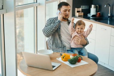 Ev elbiseli bir adam telefonda konuşuyor, kaprisli bir bebek kollarında oturuyor.