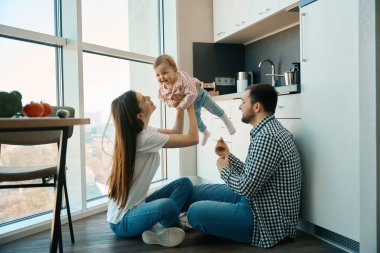 Genç ebeveynler mutfakta küçük bir çocukla bebek bakıcılığı yapıyor, aile manzara penceresinin yanında yer alıyor.