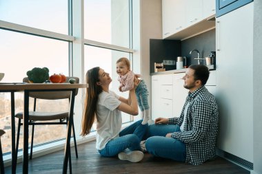 Bakıcı ebeveynler evde küçük bir çocukla mutfakta oynarlar, aile manzara penceresinde yer alır.