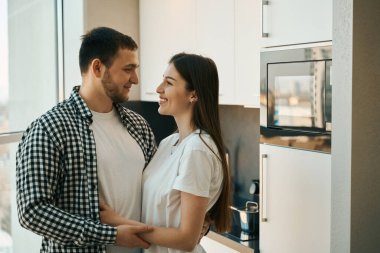 Sevimli genç eşler dikiliyor, kucaklıyor, pencerenin önünde rahat kıyafetler giyen erkekler.