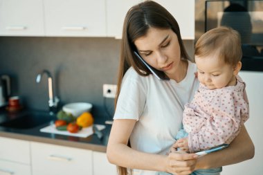 Ellerinde termometre tutan ve telefonla konuşan endişeli bir kadın. Kollarında sevimli küçük bir bebek var.