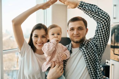 Parlak mutfakta ebeveynlerin kollarında küçük bir çocuk, anne ve baba evde bebekle oynuyorlar.