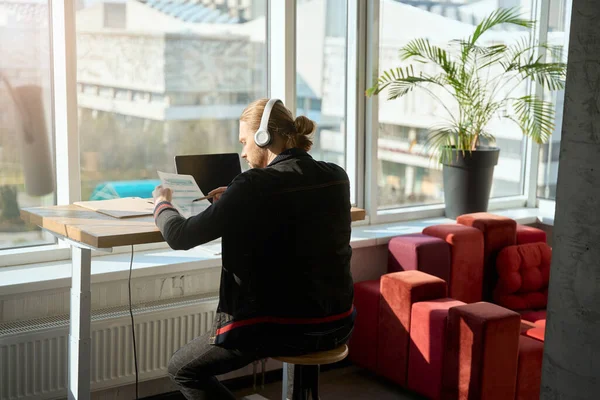 同僚のスペースでノートを閲覧しながら椅子に座ってノートパソコンで作業しているヘッドフォンの白人男性 — ストック写真