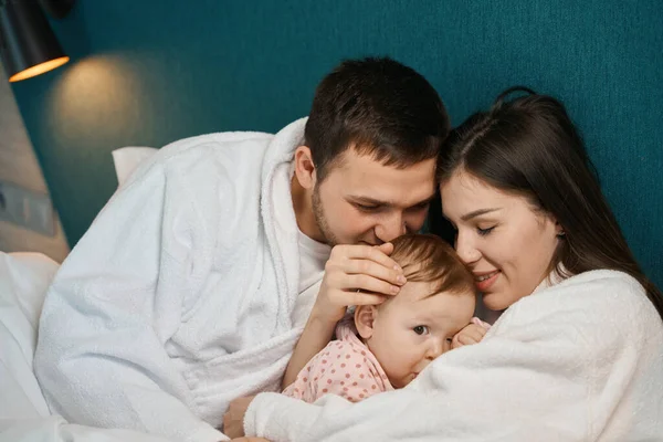 可爱的婴儿在妈妈的怀里躺在父母的床上 爸爸吻着孩子的头顶 — 图库照片
