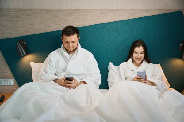 这对年轻夫妇躺在柔软的床上 手里拿着手机 心情很好 — 图库照片
