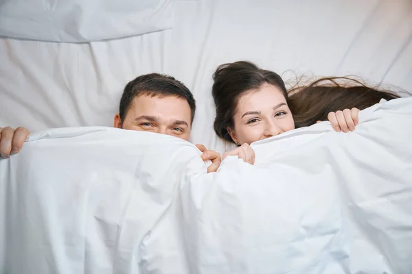 大きなベッドの上のカバーの下に笑顔の夫と妻 彼らはカバーの下からかわいい見えます — ストック写真
