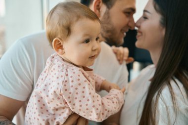 Babalarının kollarında tombul yanakları olan küçük güzel bir kız. Ebeveynler birbirlerine sevgiyle bakar.