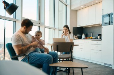 Bakıcı baba, küçük kızına mutfakta bakıyor. Yanında da dizüstü bilgisayarı olan bir anne.