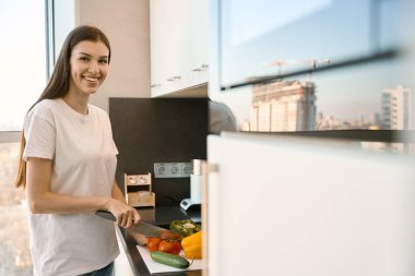 Neşeli kadın salata için parlak bir mutfakta sebze keser, bir kadın manzaranın karşısında durur.