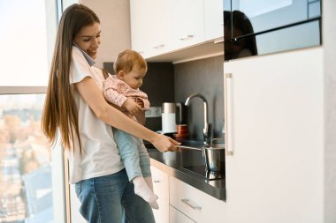 Neşeli genç anne kucağında küçük yardımcısıyla bebek maması hazırlıyor. Kadın cep telefonuyla konuşuyor.