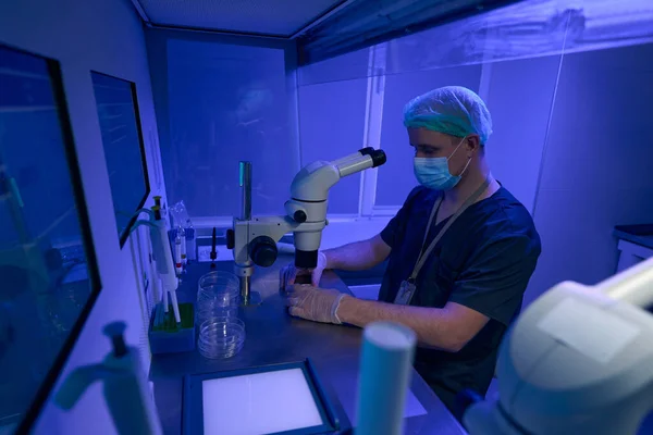 实验室显微镜下 穿着无菌制服 戴防护面罩 戴手套 戴帽子的男子用紫外线进行研究 — 图库照片