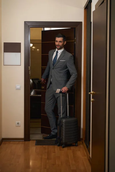 旅行用のスーツケースを着た男がホテルの部屋を出ると 彼はビジネススーツに身を包んだ — ストック写真