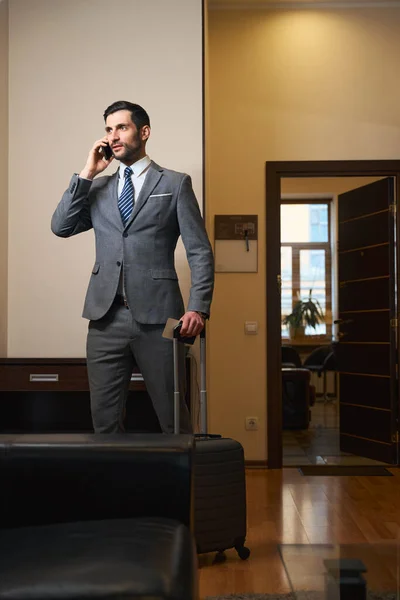 Hotel guest in a business suit communicates on a mobile phone, a man has a travel suitcase