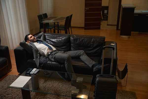 在酒店房间里的一个舒适的沙发上 一个旅行的男人在路上休息 旁边是一个旅行的行李箱 — 图库照片