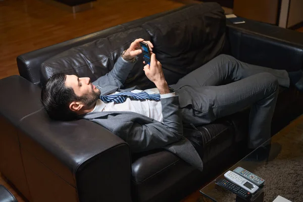 Man rests on a leather sofa in a hotel room, he uses a mobile phone