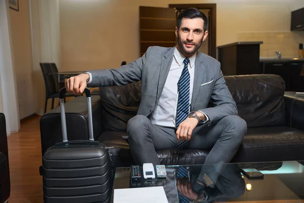 Satisfied man in a business suit sits on a cozy sofa, he has an expensive watch on his hand