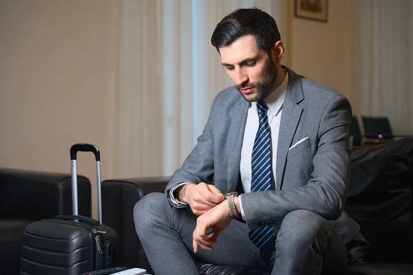 Hotel guest sits on a leather sofa and looks at his wristwatch, next to a travel suitcase