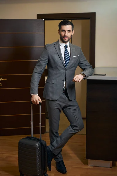 Middle-aged hotel guest stands with a travel suitcase at the counter, a man in a business suit