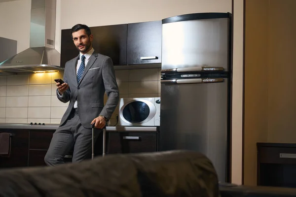 Smiling man with mobile phone stands in the kitchen area of hotel room, next to travel suitcase