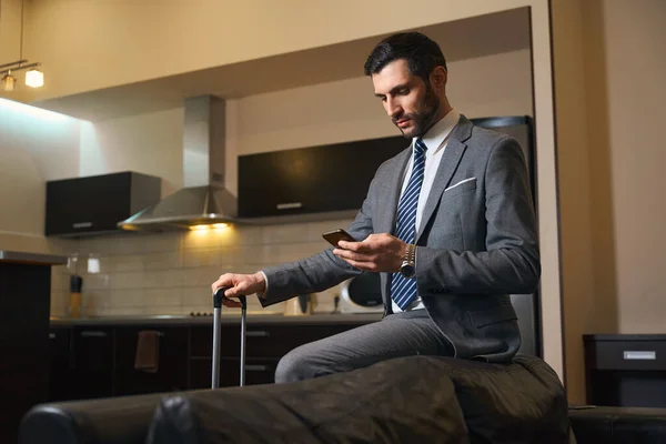 革のソファの後ろに携帯電話を持つ男 近くの旅行スーツケーススタンド — ストック写真