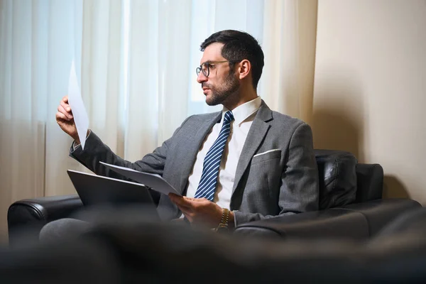 穿着西装和眼镜的男人正在休闲区学习工作文件 他坐在皮椅上 — 图库照片