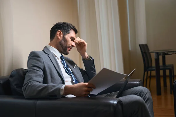 ビジネススーツを着た疲れた男が革の椅子に座って 彼は作業文書とラップトップを持っています — ストック写真