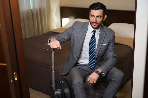 旅のスーツケースを着た男がホテルの部屋の大きなベッドの上に座って 彼はビジネススーツを着ています — ストック写真