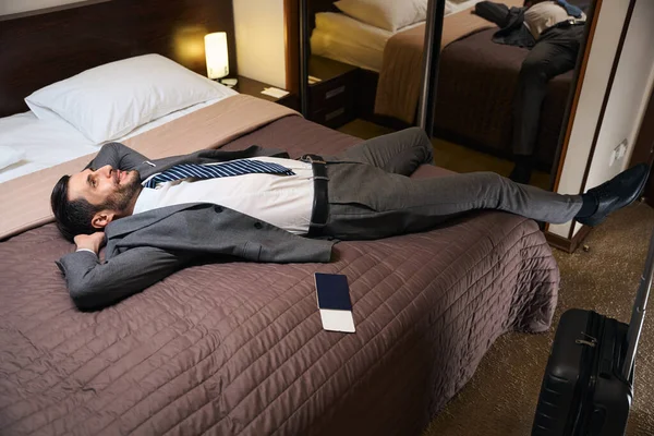 男人在旅馆房间的一张大床上躺在路上 旁边是旅行箱和护照 — 图库照片