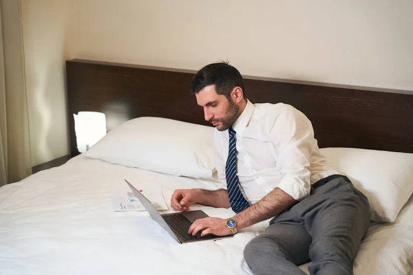 Unshaven middle-aged brunette reclining on the bed with working documents and a laptop