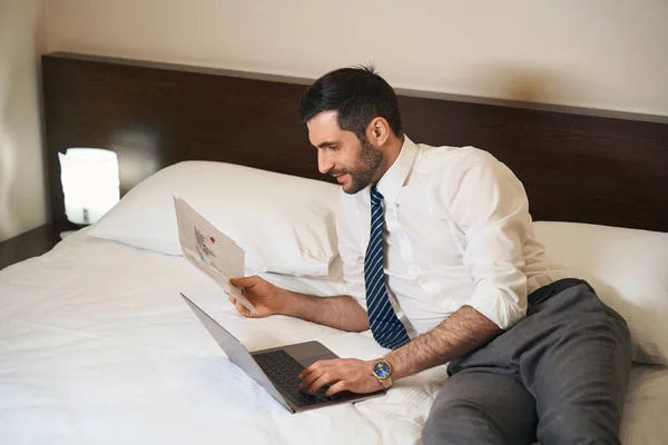 ビジネス服でUnshavenブルネットワーキングドキュメントとラップトップでベッドの上にリクライニング — ストック写真