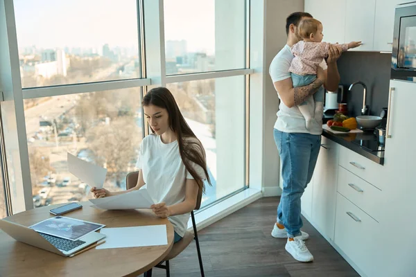 年轻女人在厨房的全景窗前远程工作 旁边是抱着孩子的丈夫 — 图库照片