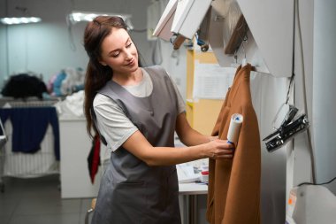 Gülümseyen çamaşırcı kadın, yapışkan iplik çıkarıcı ve profesyonel kuru temizleme servisiyle hassas kumaştan paltoyu dikkatlice temizliyor.