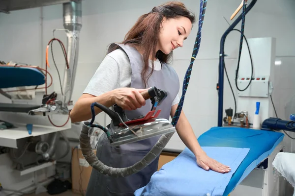 Skilled woman laundry worker getting rid of wrinkles on shirt with hot industrial iron, professional appliances and facilities