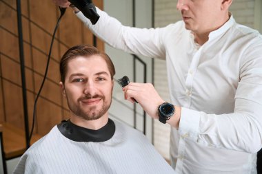 Koruyucu pelerinli adam berber koltuğunda oturuyor, usta müşterinin saçını şekillendiriyor.