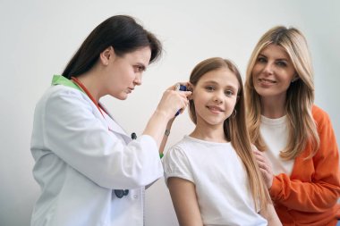 Konsantre kadın laringolojist. Özel otoskopu olan gülümseyen küçük bir hastanın kulağını inceliyor. Küçük kız acısız sağlık kontrolünden memnun.