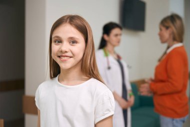 Çocuk hastalıkları kliniğinin mutlu hastası kameraya bakıyor ve gülümsüyor. Annem de sağlık testinin sonuçlarını doktorla tartışıyor.