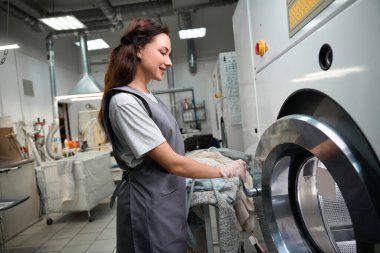 Gülümseyen kadın çamaşır teknisyeni kirleri temizlemek ve lekeleri kaldırmak için kimyasal çözücü ile kuru temizleme makinesine çamaşır koyuyor.