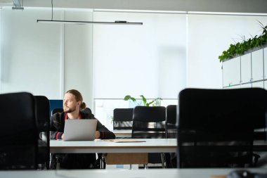 Genç adam büyük bir konferans masasında oturuyor, odanın içinde minimalist bir iç mekan var.