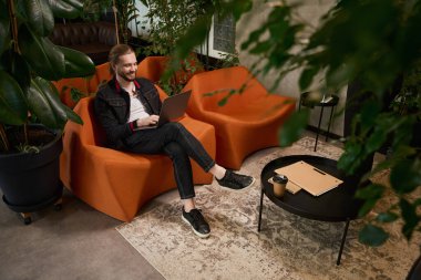 Sakallı erkek, dinlenme odasında dizüstü bilgisayarla birlikte, sehpanın üzerinde dosyalar ve kahve var.