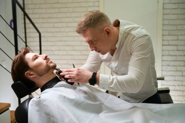 Berber dükkanında sakal tıraşı olan genç bir adam, sakal kesme makinesi kullanan bir uzman.