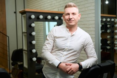 Açık renk gömlekli berber aynalı ve mükemmel ışıklandırmalı iş yerinde duruyor. Adamın akıllı bir saati var.